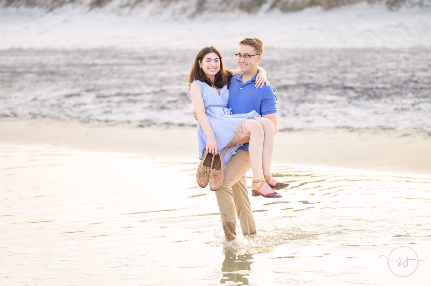 Man in a blue shirt carrying his new fiancée across a shallow tide pool at Huntington Beach State Park