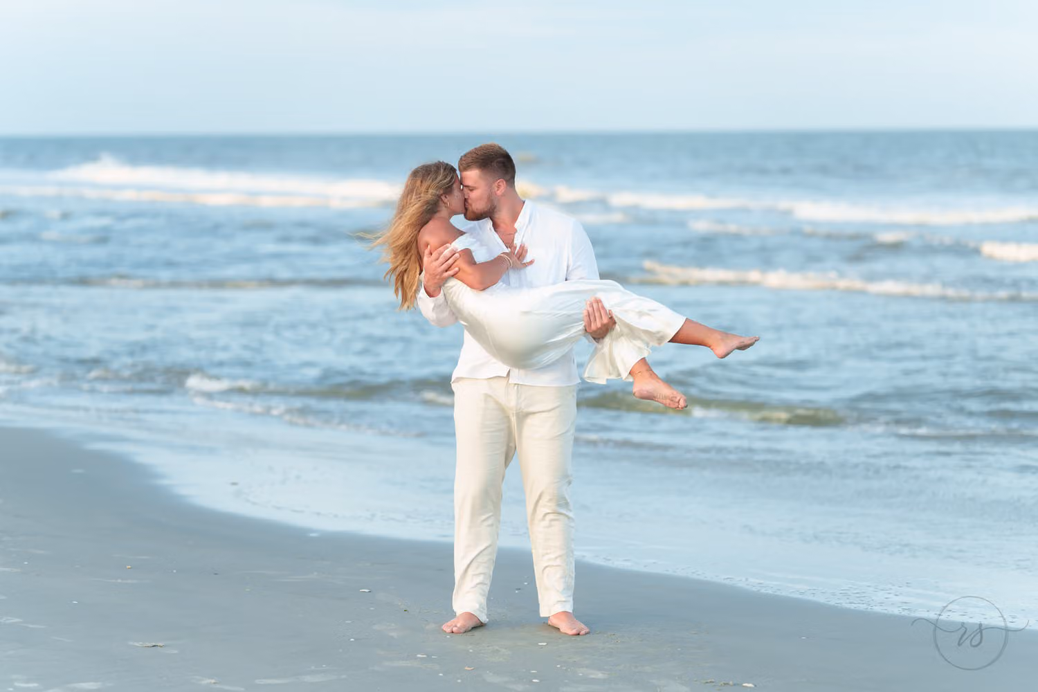 Man in white carrying his new fiancée in his arms as they kiss along the shoreline in Pawleys Island