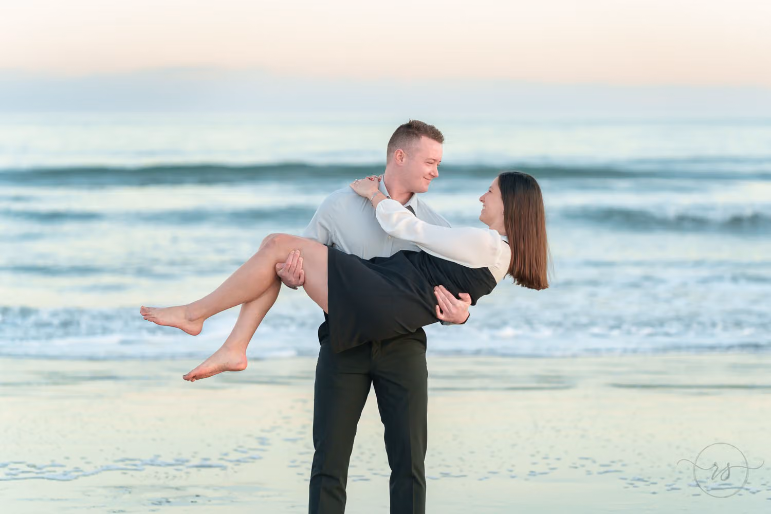 Man carrying his fiancée in his arms by the gentle waves in soft evening light at Huntington Beach State Park