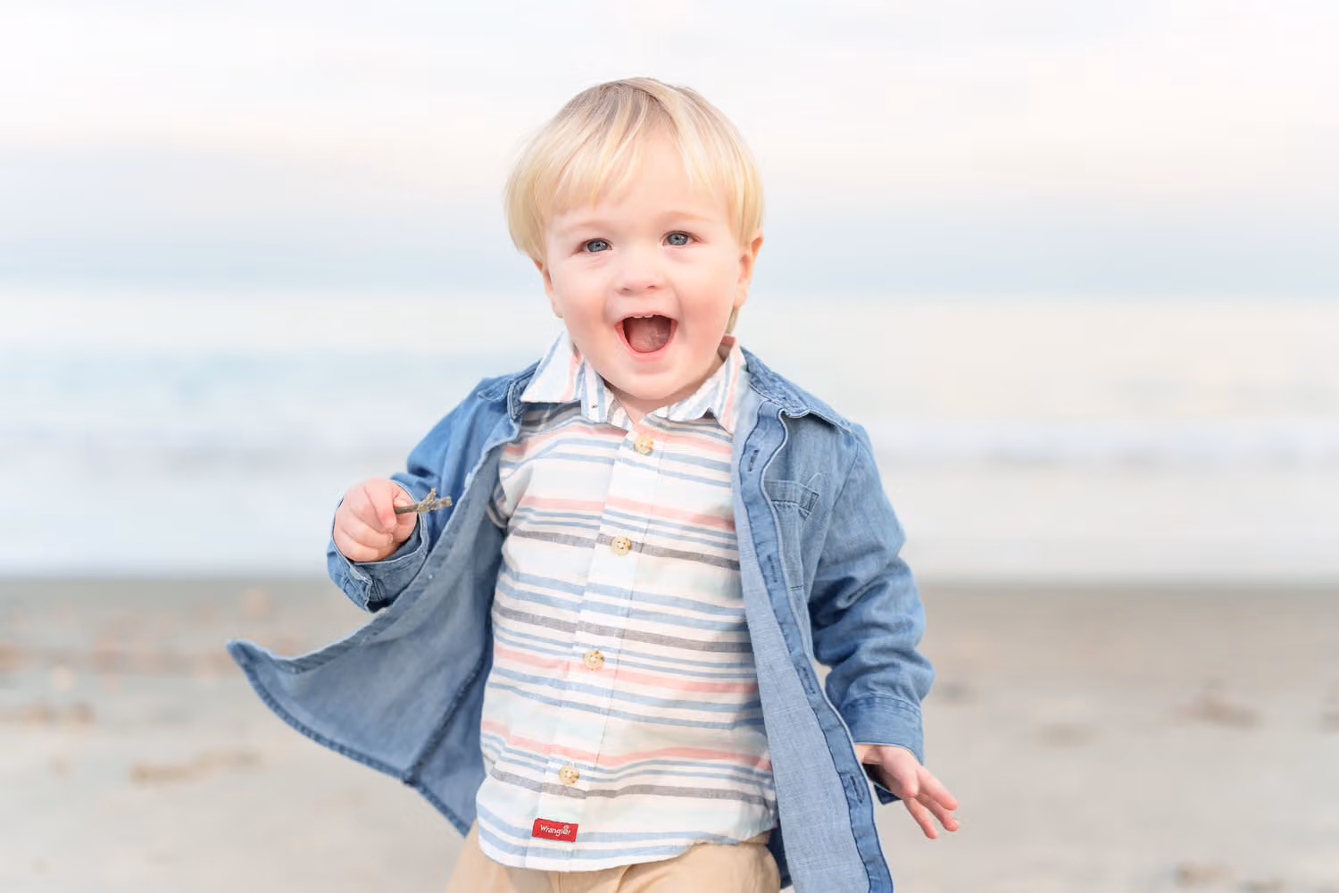 Laughing little blond boy in a denim jacket running across the sand toward the camera at Huntington Beach State Park