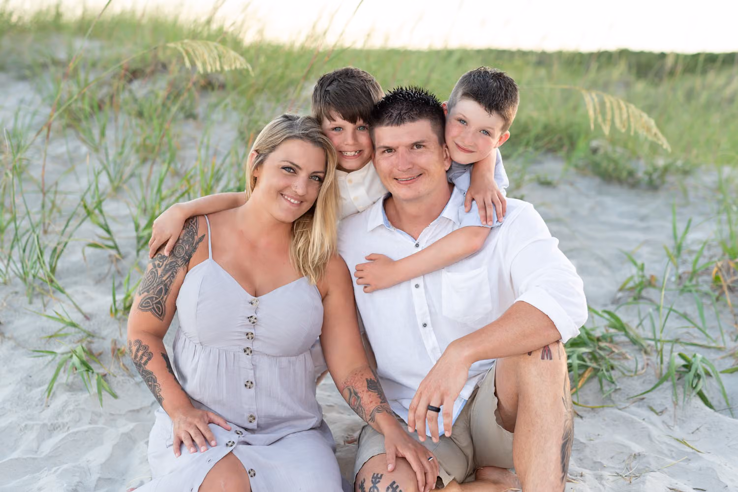 Mom and dad sitting in the sand by the dunes with their two boys hugging them from behind at Huntington Beach State Park