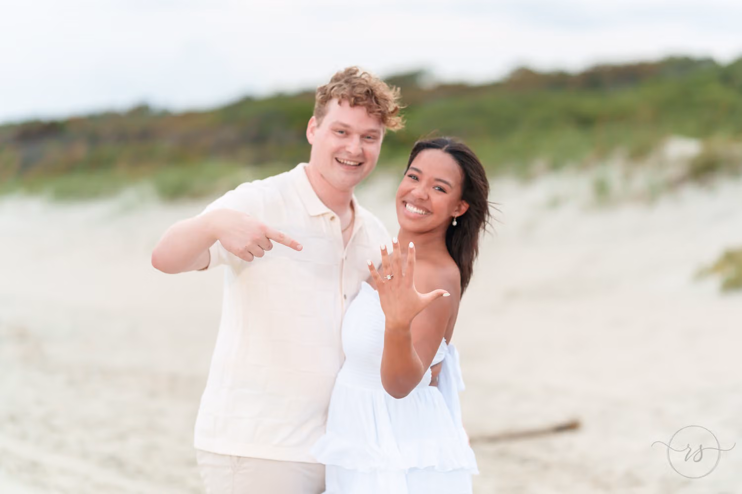 Newly engaged couple smiling and pointing at her engagement ring in front of the dunes at Myrtle Beach State Park