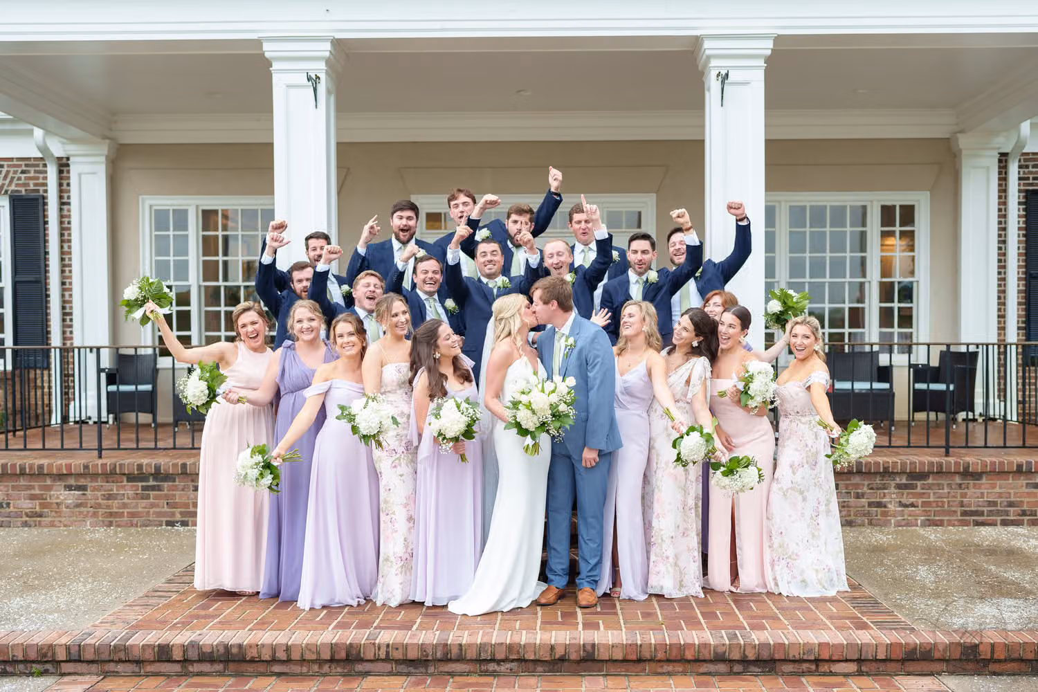 Big wedding party in pastel dresses cheering around the couple on the brick steps behind the clubhouse at Pawleys Plantation