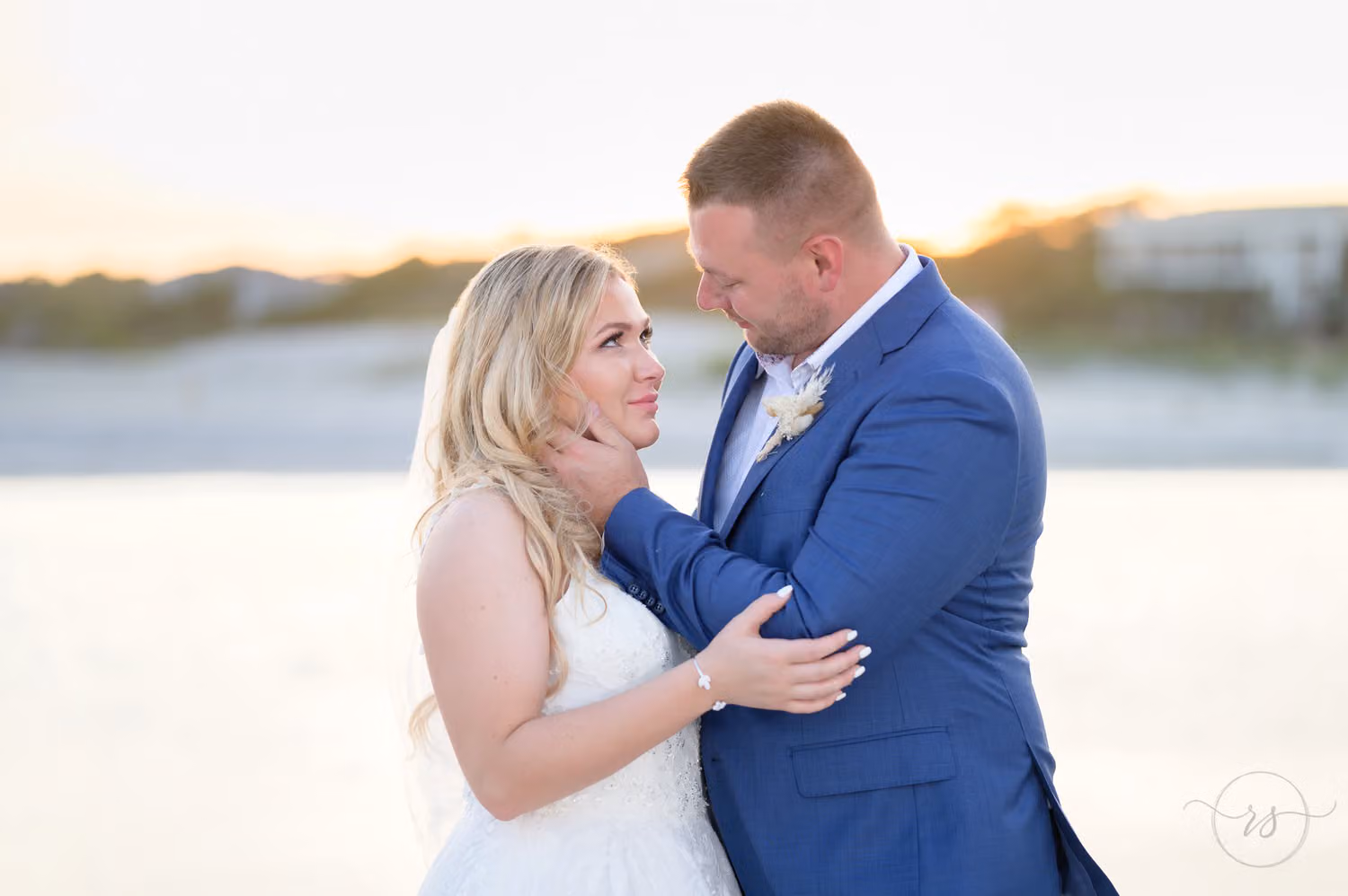 Close-up of the groom in a blue suit pulling the bride close by the water at sunset at North Beach Resort & Villas