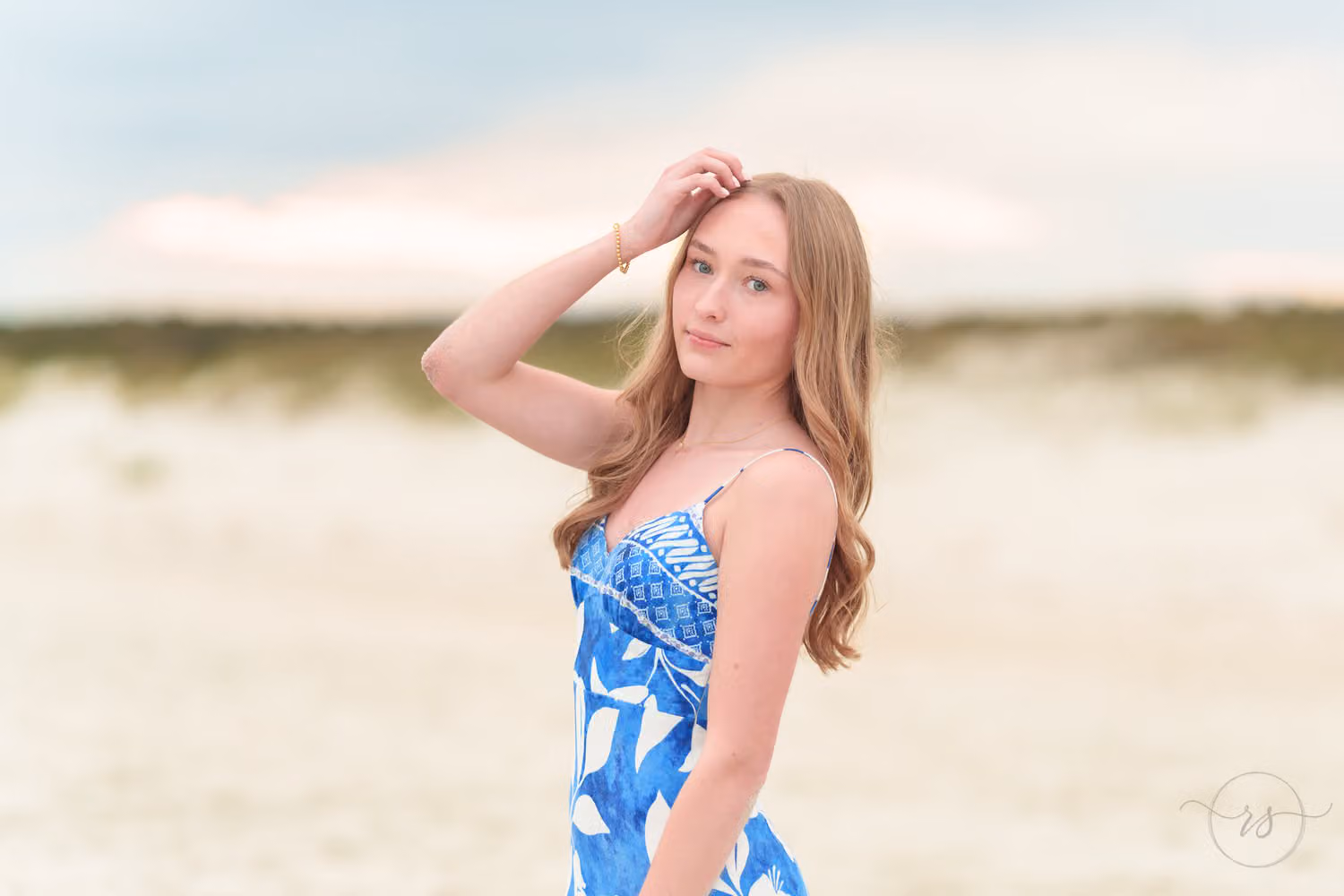 Close portrait of a senior girl in a blue dress, hand in her hair, with a soft sunset behind at Huntington Beach State Park