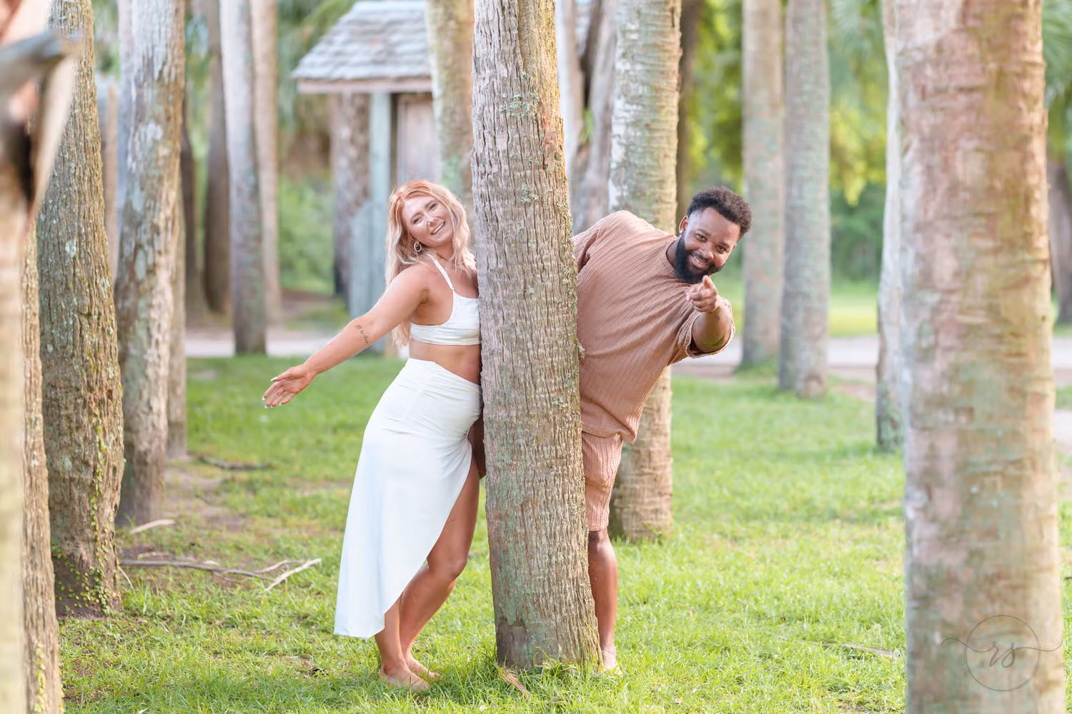 Couple playfully peeking around palm trunks in a grassy grove at Huntington Beach State Park