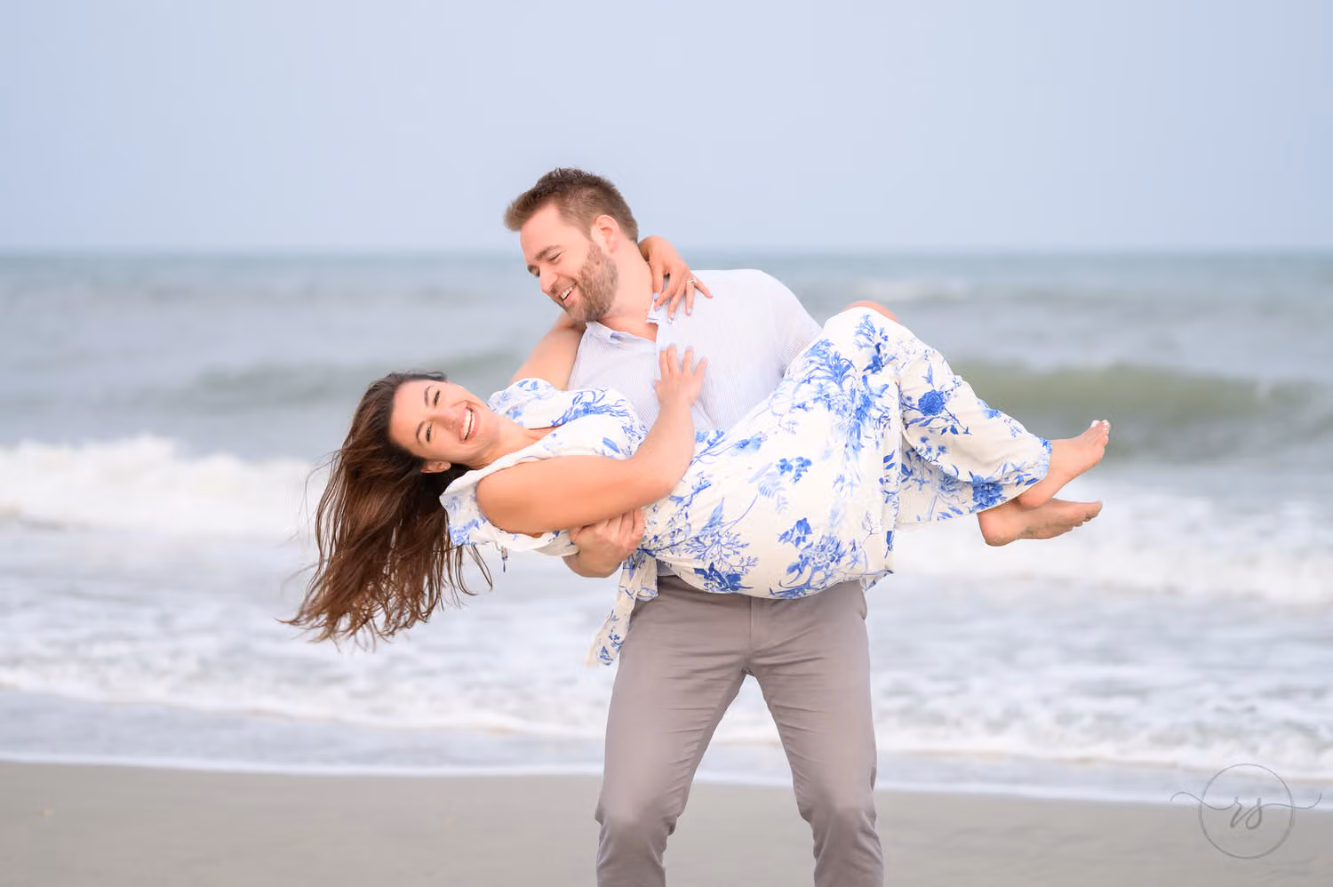 Man carrying his laughing fiancée in a blue floral dress as she leans back by the ocean at Huntington Beach State Park