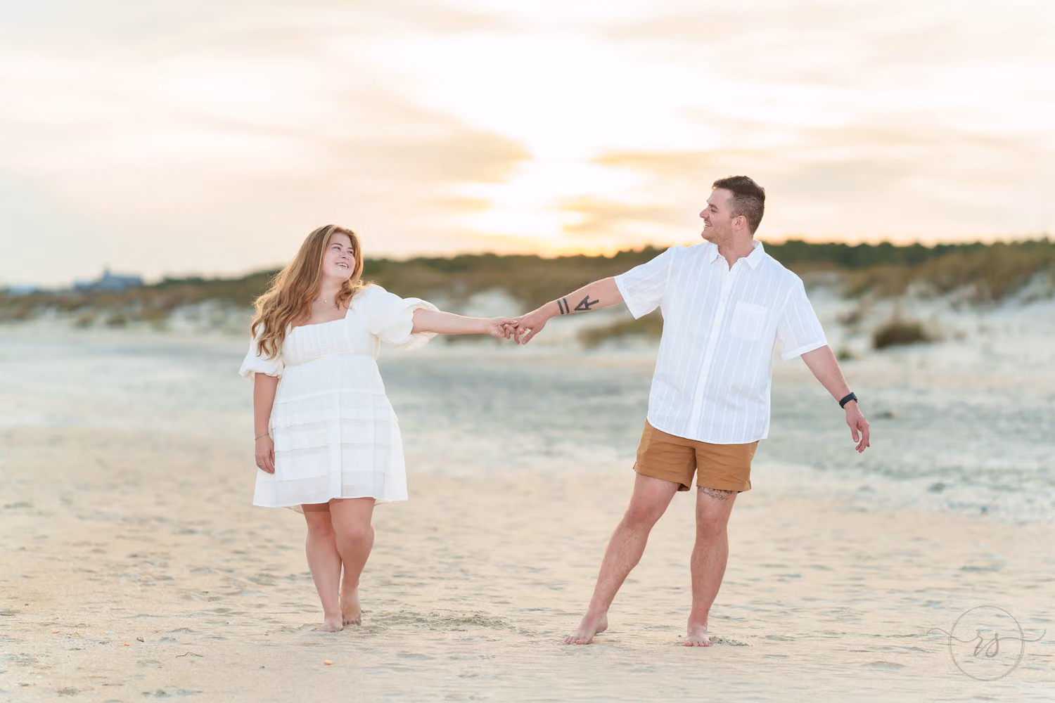 Couple in white reaching out to hold hands on the beach with the sun setting behind them at Huntington Beach State Park