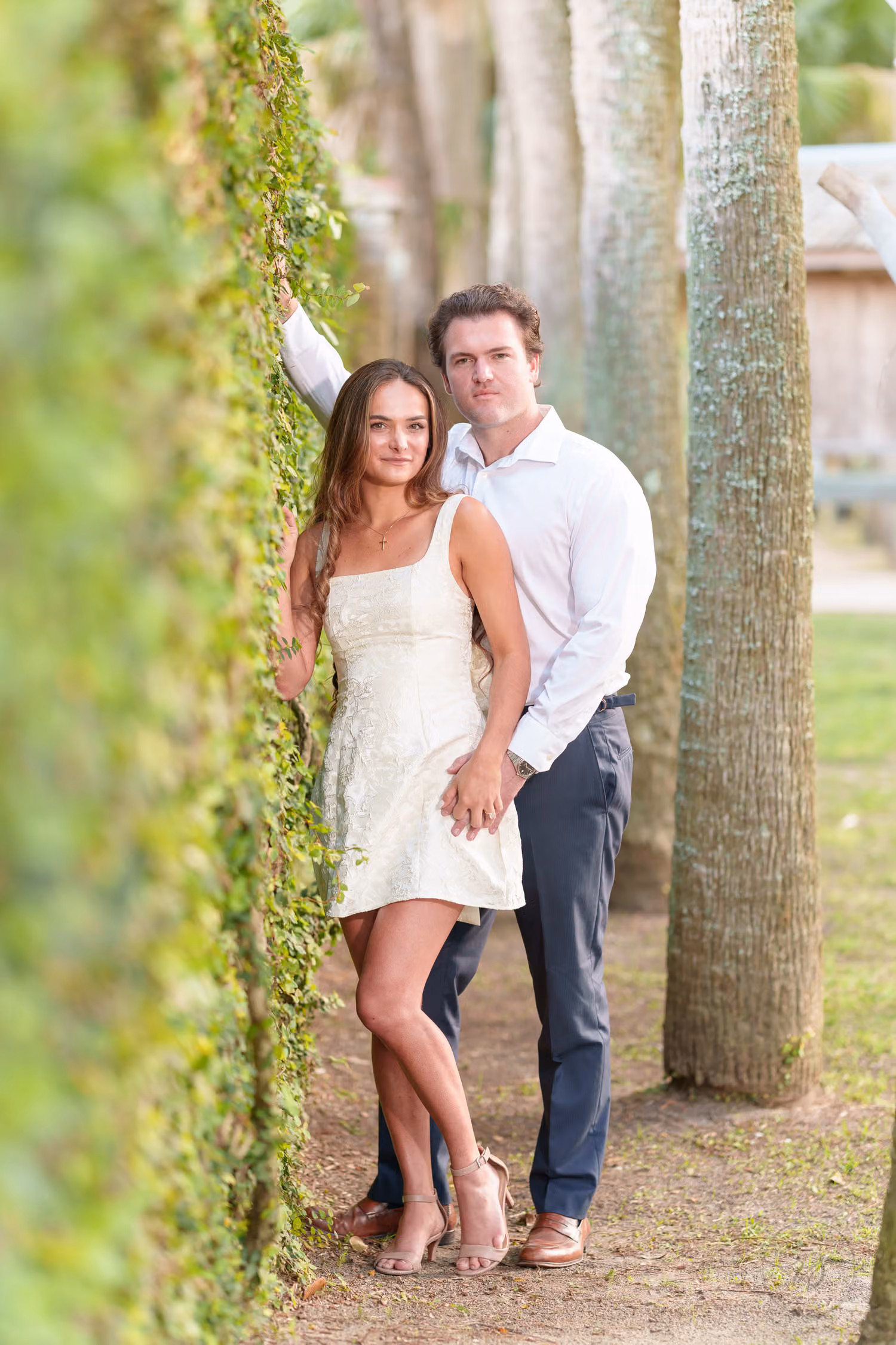 Couple standing close against an ivy-covered wall beside a row of tall palm trunks at Huntington Beach State Park