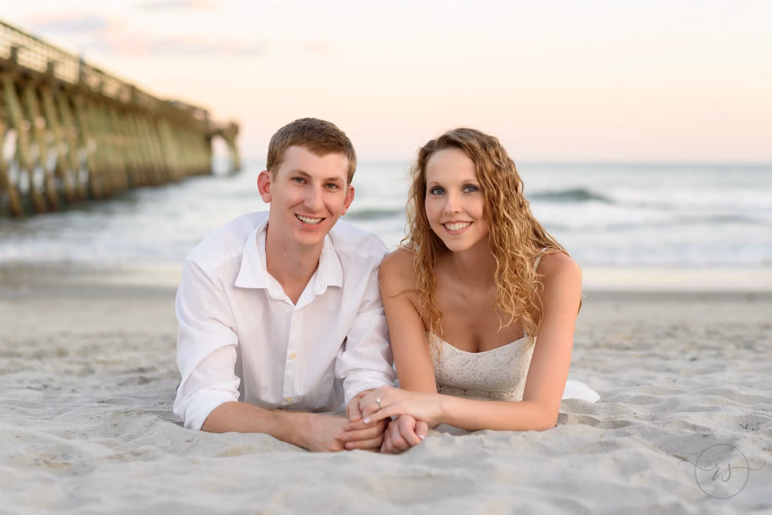 Couple lying side by side in the sand, smiling, with the pier stretching out behind them at Myrtle Beach State Park