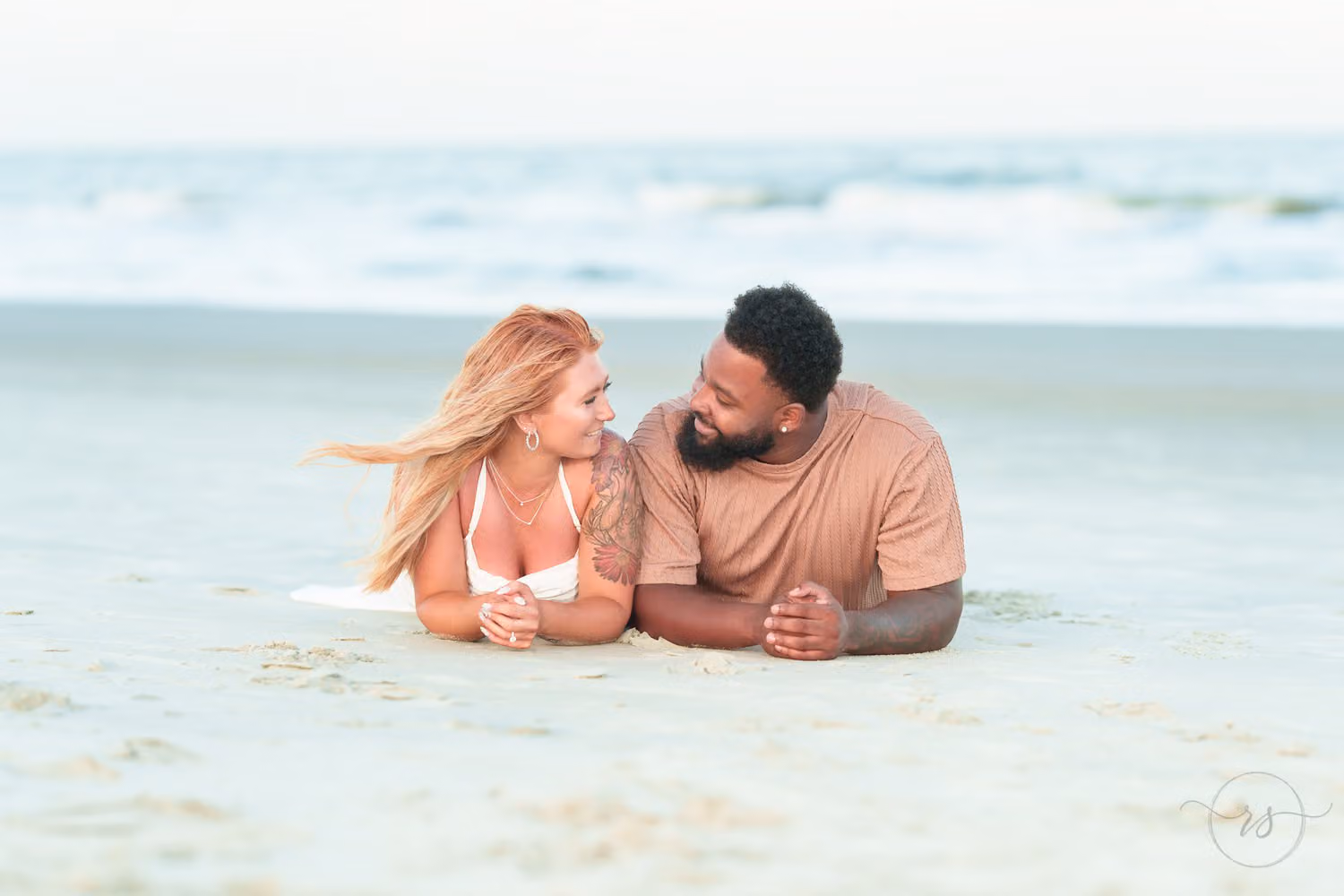 Couple lying side by side on their stomachs in the sand, smiling at each other at Huntington Beach State Park