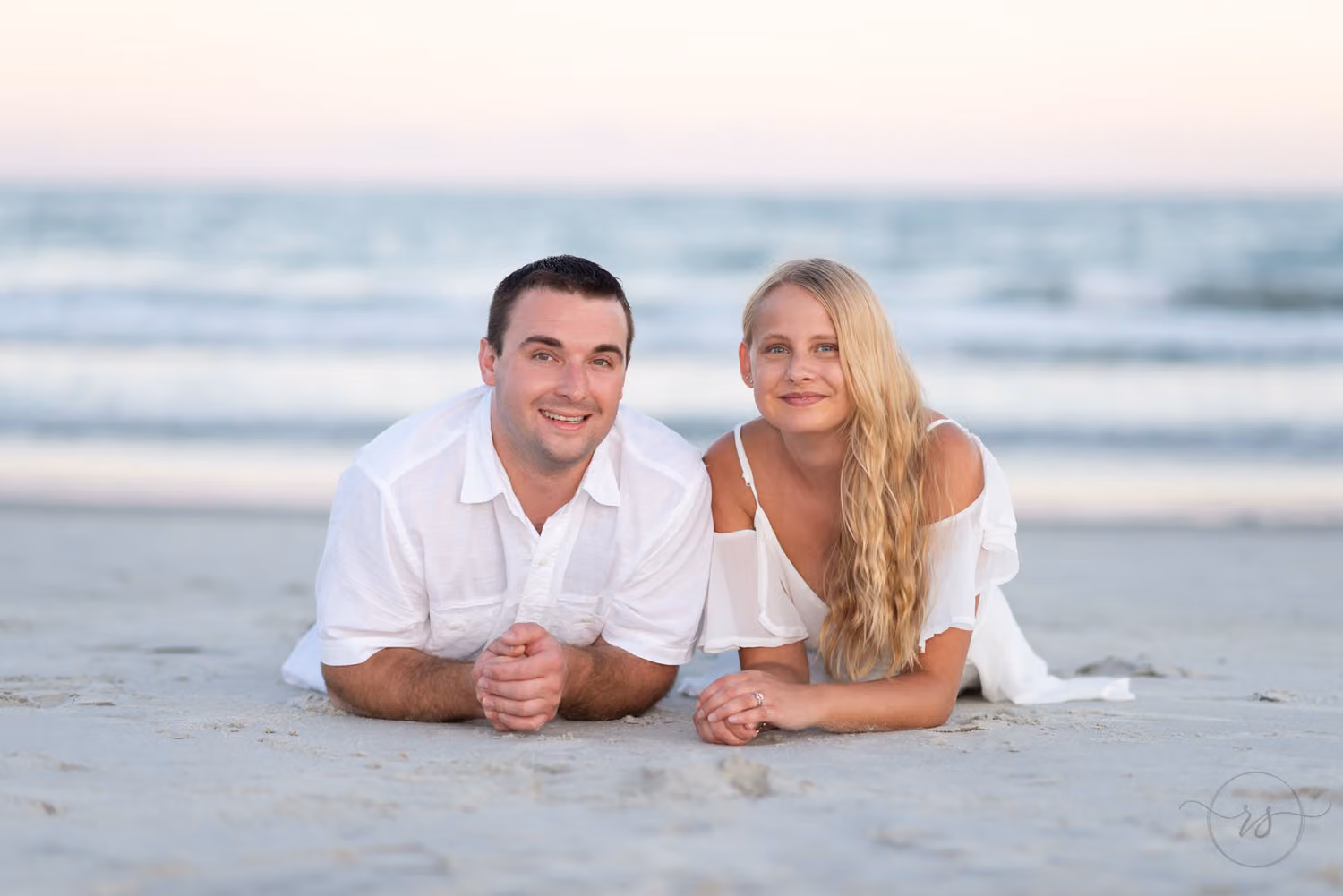 Couple lying on their stomachs in the sand smiling at the camera with the ocean behind them at Huntington Beach State Park