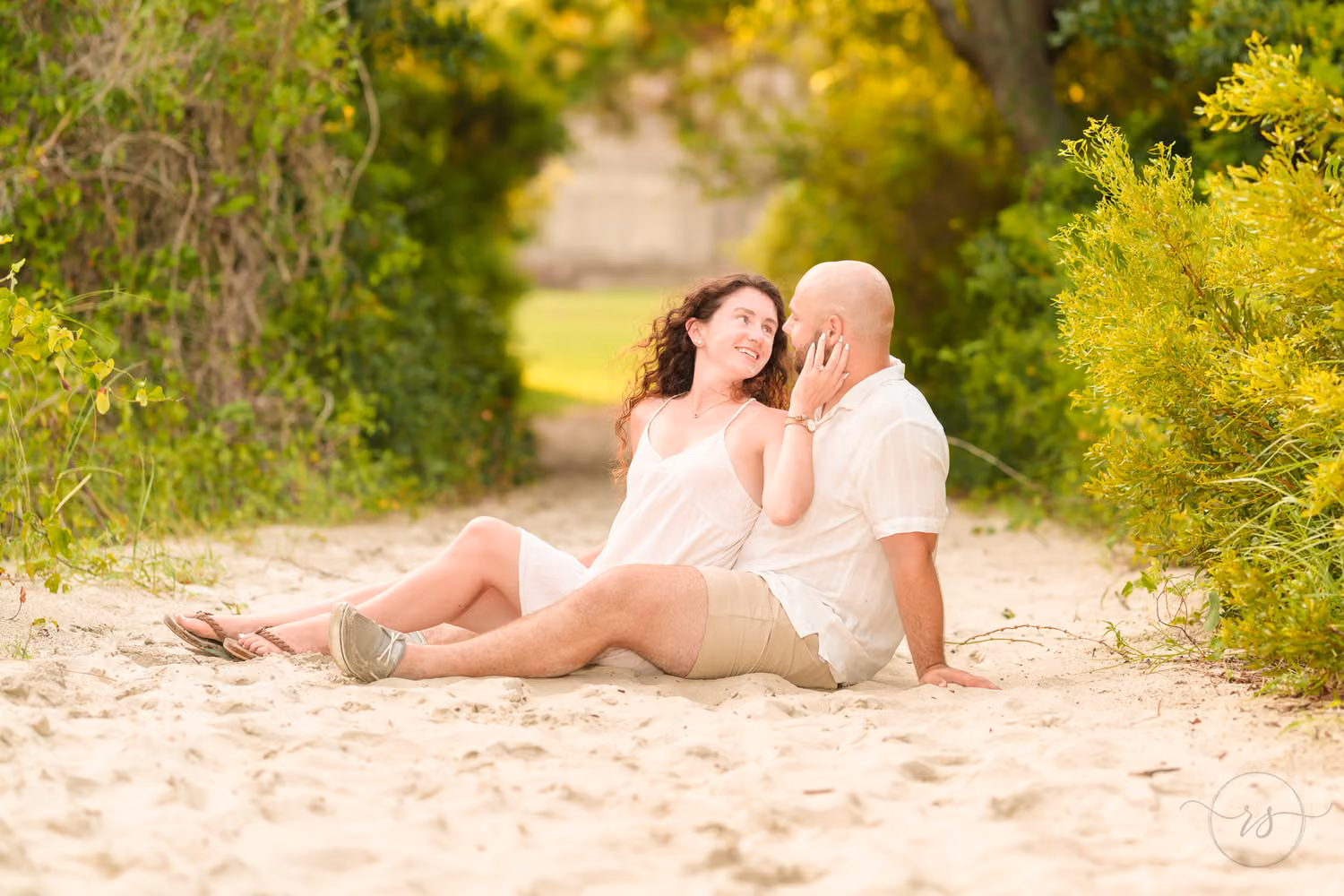 Couple sitting in the sand on a beach path framed by green bushes at Huntington Beach State Park