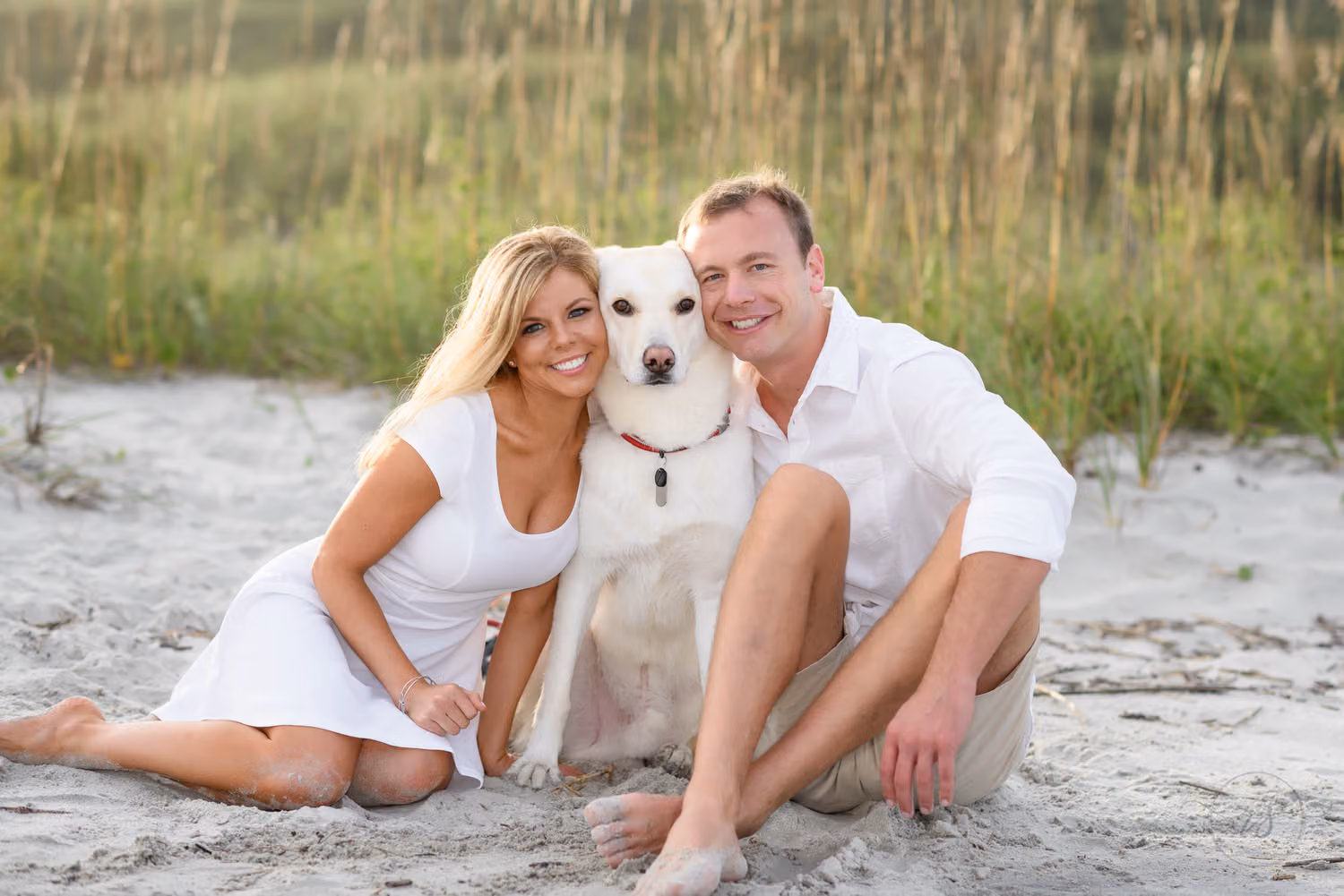 Couple sitting in the sand with their white dog between them in front of the dunes at Myrtle Beach State Park