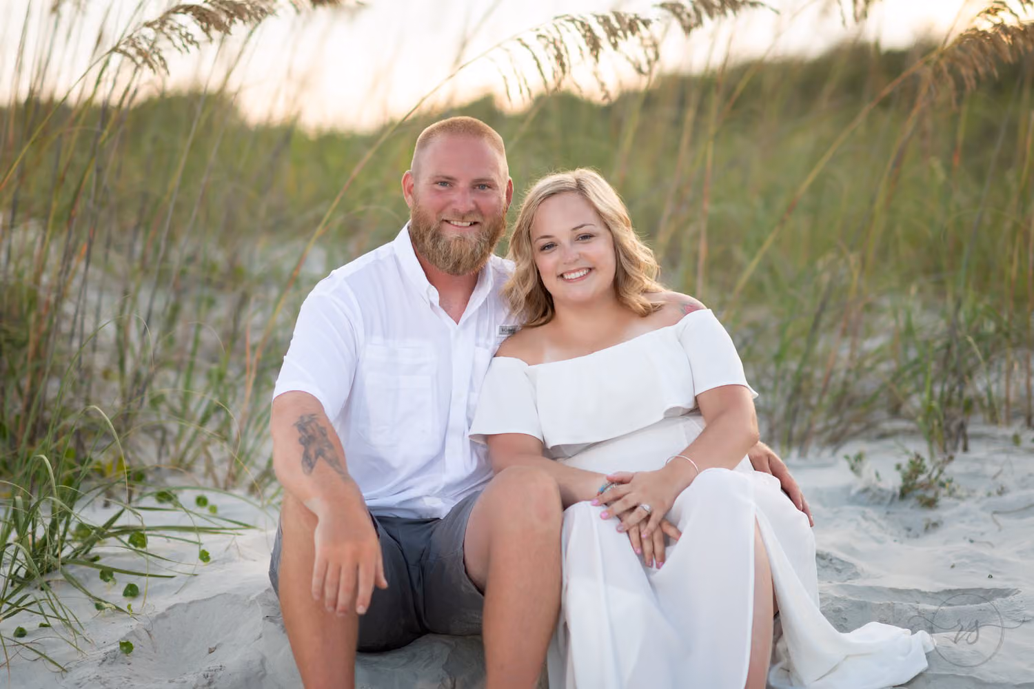 Couple dressed in white sitting together in the sand in front of sea oats at sunset