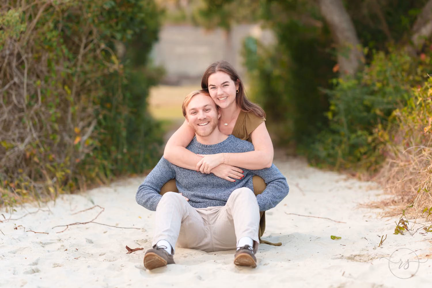 Woman hugging her fiancé from behind as they sit on a sandy path framed by green bushes at Huntington Beach State Park
