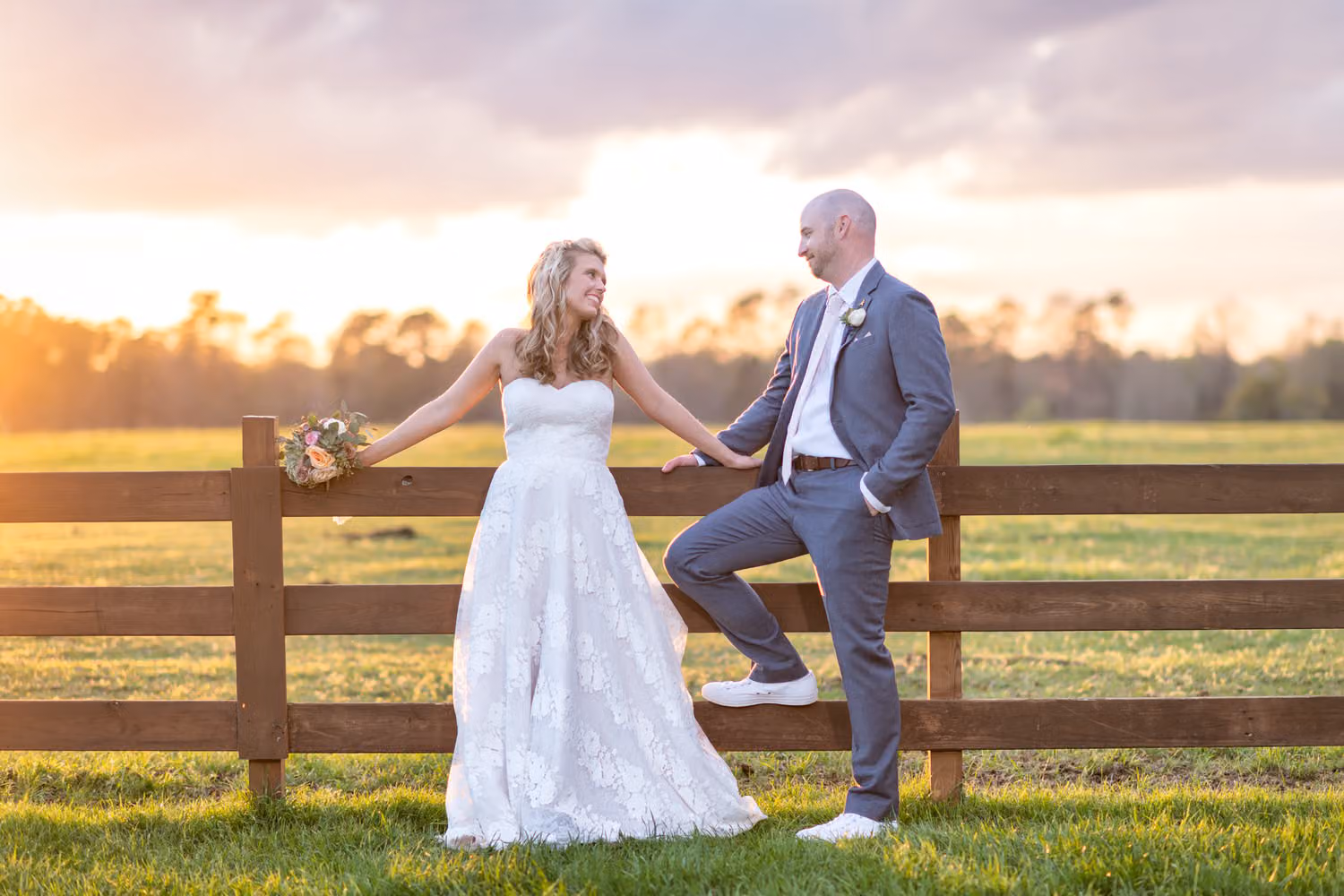 Bride and groom holding hands as they lean on a wooden fence with one foot up at sunset at Wildhorse at Parker Farms