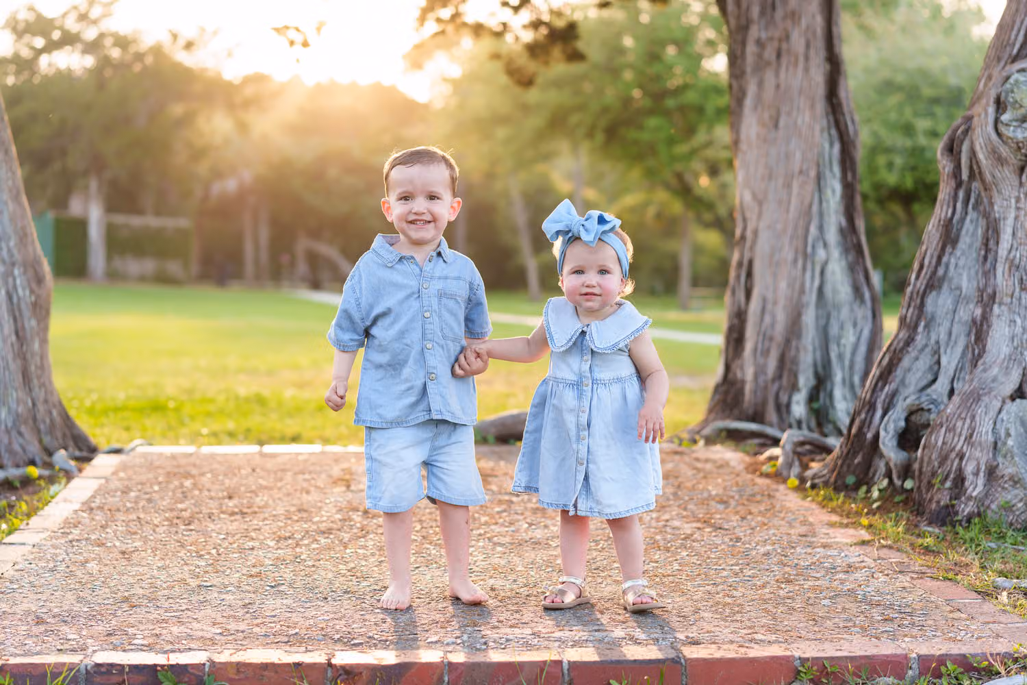 Toddler brother and sister in blue holding hands on a sandy path between oak trunks at sunset in Pawleys Island