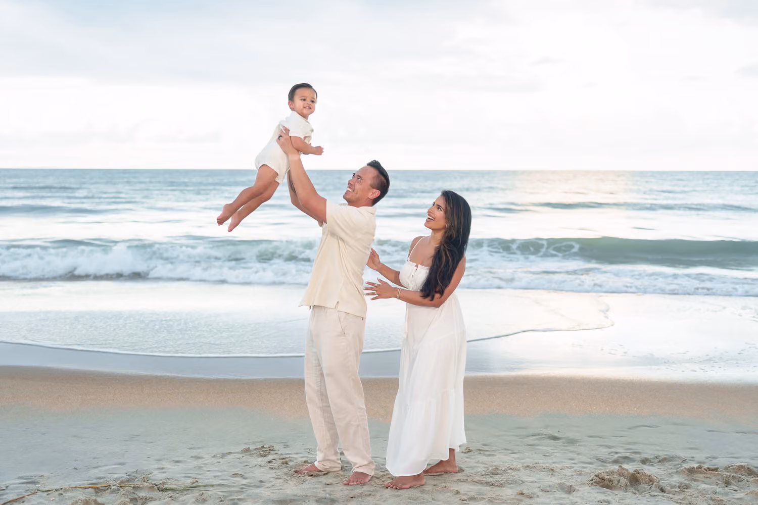 Dad lifting his little boy high over his head as mom smiles beside him by the ocean at Huntington Beach State Park