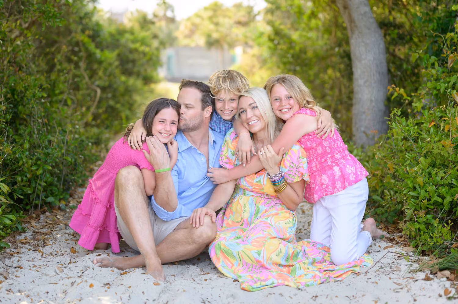 Family of four sitting close in the sand on the tree-lined beach path, all hugging and smiling at Huntington Beach State Park