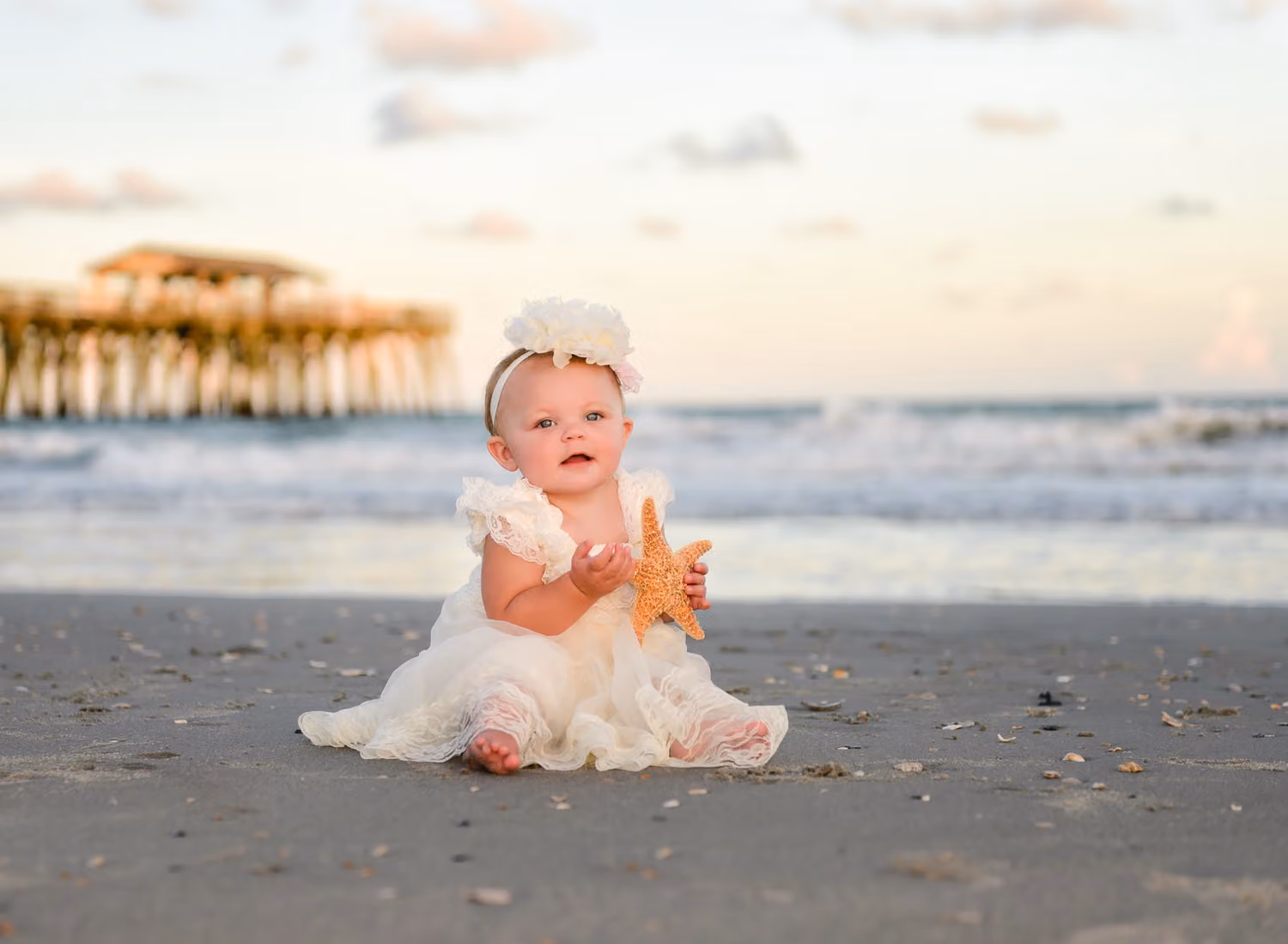 Baby girl in a white dress and flower headband holding a starfish on the sand with the pier behind at Myrtle Beach State Park