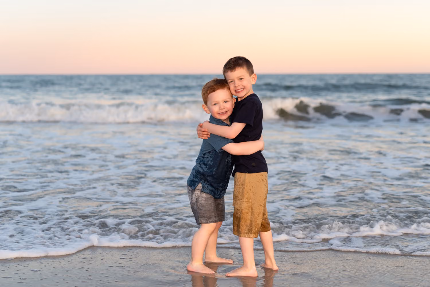 Two little brothers hugging at the water's edge with waves rolling in behind them at sunset
