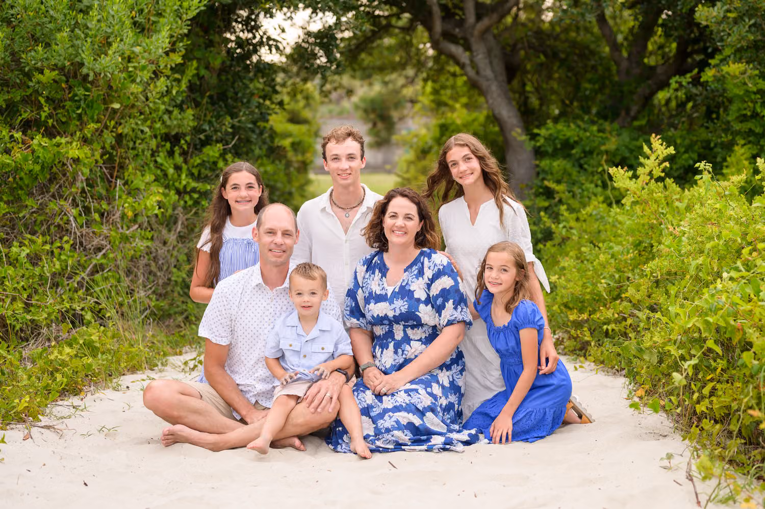 Family of seven in blue and white sitting together on the sandy path under the trees at Huntington Beach State Park