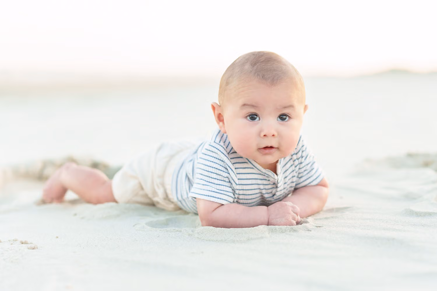 Wide-eyed baby boy lying on his tummy in the sand looking up at the camera at Huntington Beach State Park