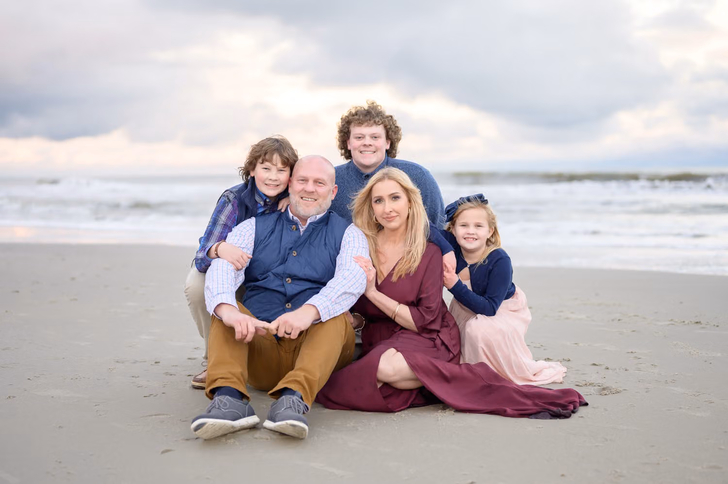 Family of five snuggled together on the sand with the ocean and cloudy sky behind them at Huntington Beach State Park