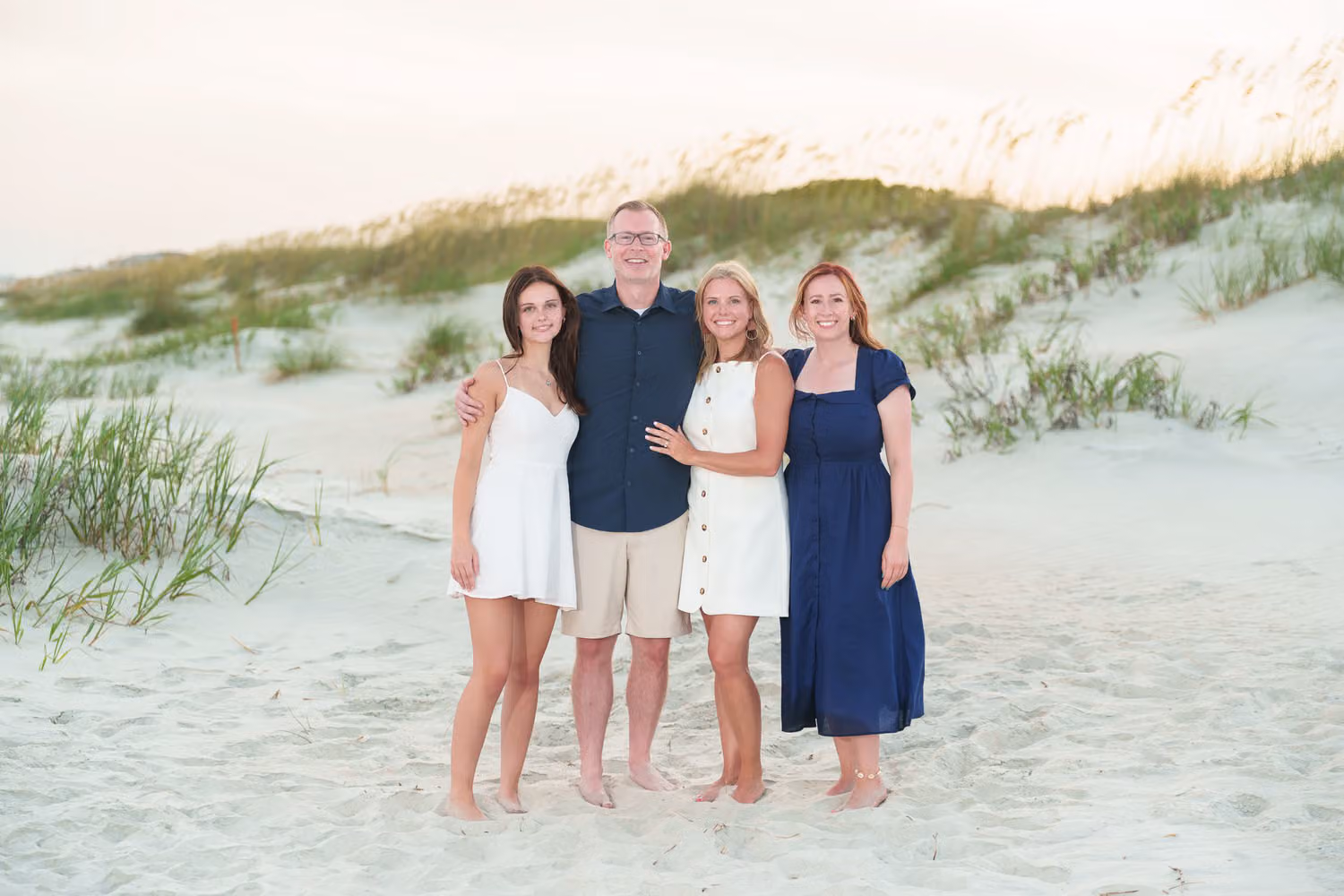 Family of four standing together on the sand in front of the grassy dunes at Huntington Beach State Park