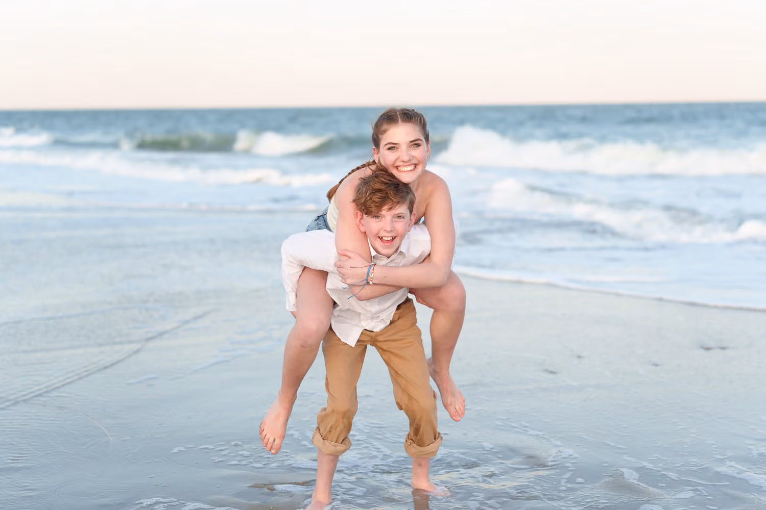 Teen boy carrying his laughing big sister piggyback in the shallow water at Huntington Beach State Park