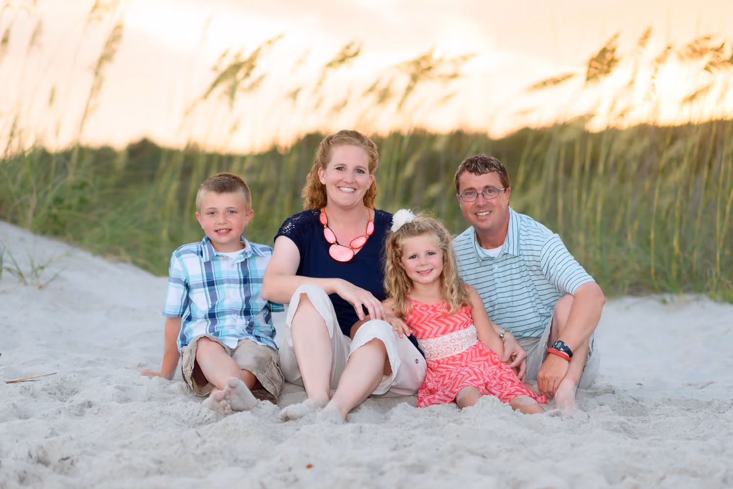 Family of four with a son and daughter sitting in the sand in front of sea oats at sunset in Myrtle Beach