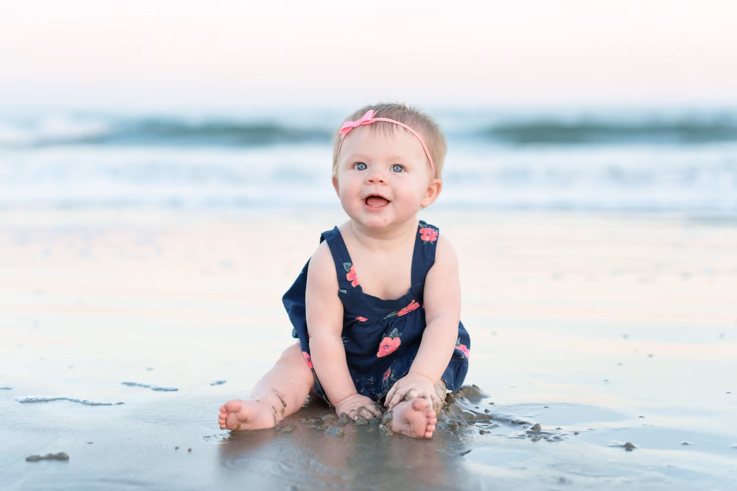 Toddler girl in a navy romper and pink bow sitting in the wet sand by the waves at Huntington Beach State Park