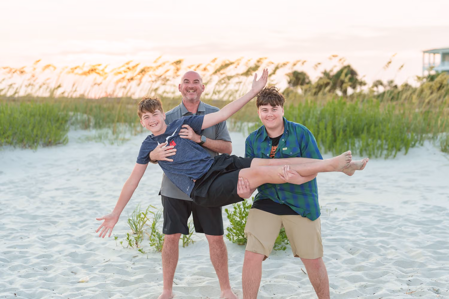 Dad holding one son sideways while his brother laughs beside them by the sea oats at Garden City Beach