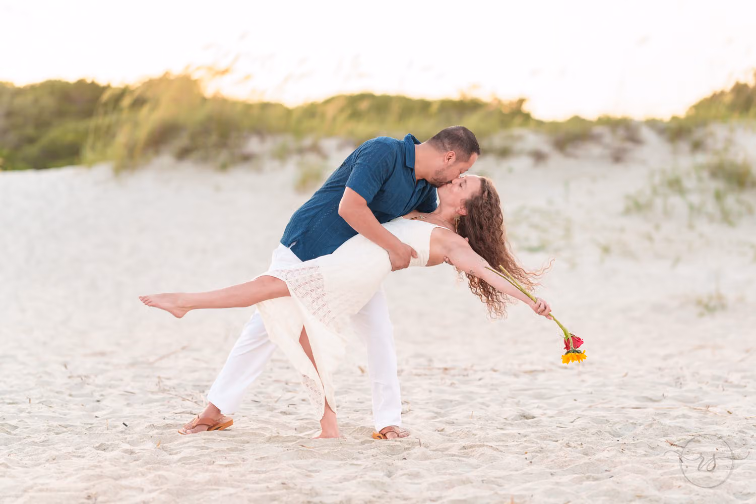 Woman holding a small bouquet as her fiancé dips her low in the sand by the dunes at Huntington Beach State Park