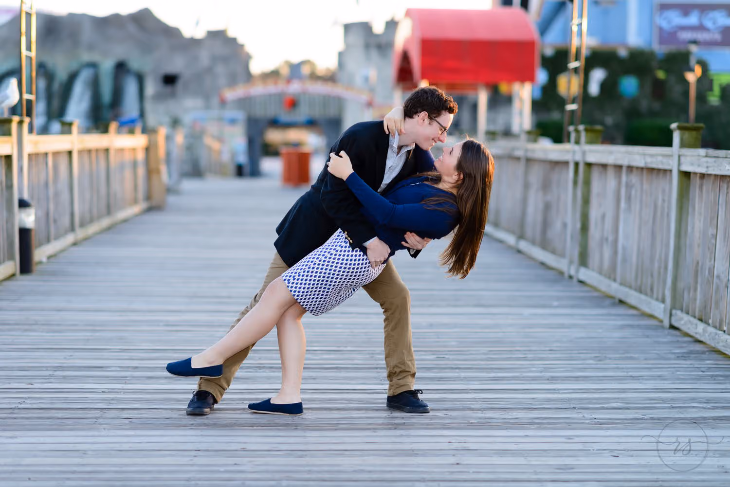Man dipping his fiancée back for a kiss on the wooden boardwalk at Broadway at the Beach