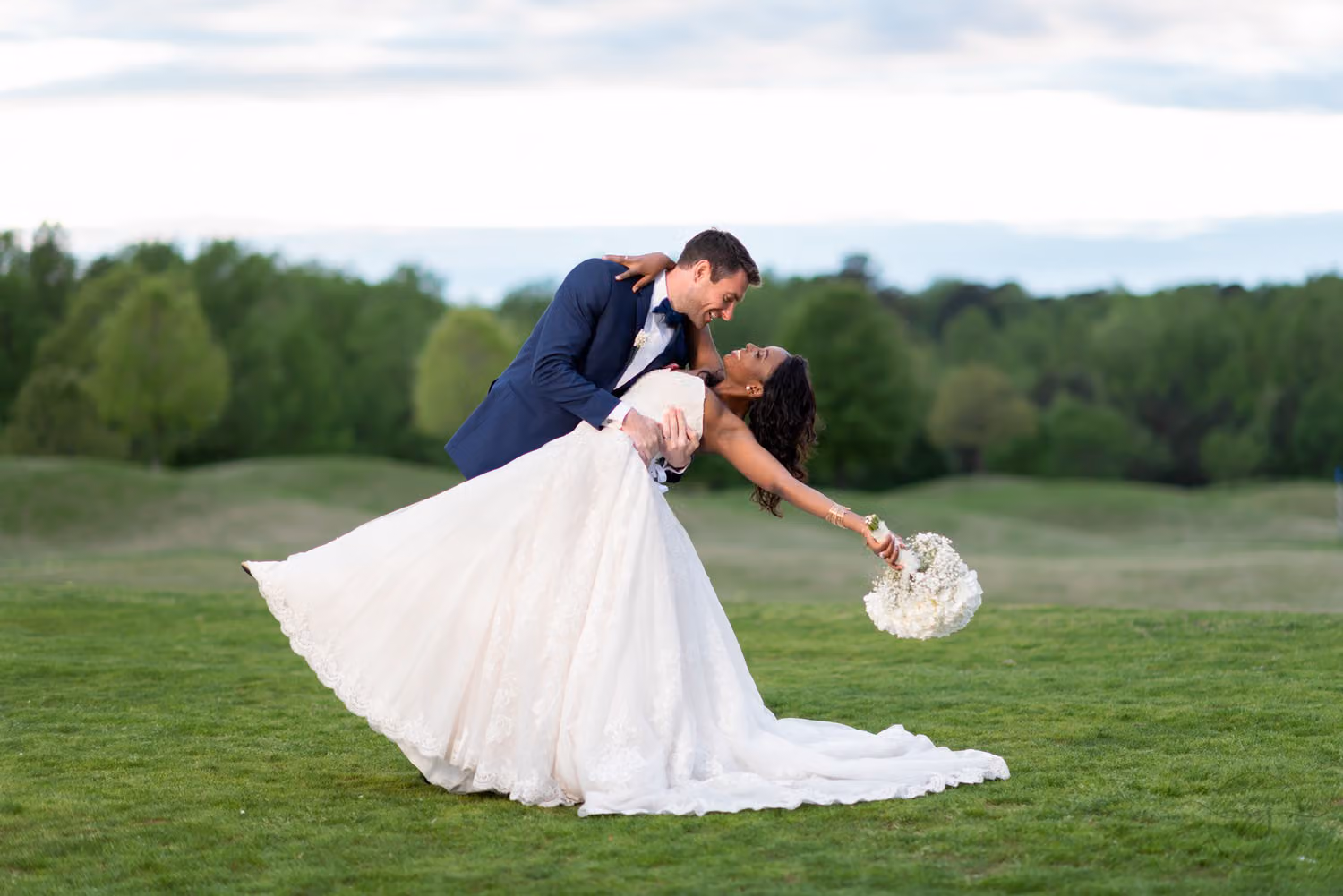 Groom in navy dipping the bride far back as she holds her bouquet out on a grassy golf course hill at River Ridge Golf Club