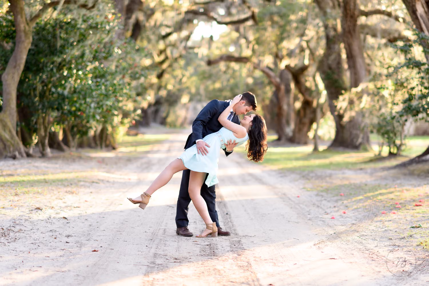 Man dipping his fiancée in a light blue dress for a kiss on a dirt road under live oaks at Mansfield Plantation
