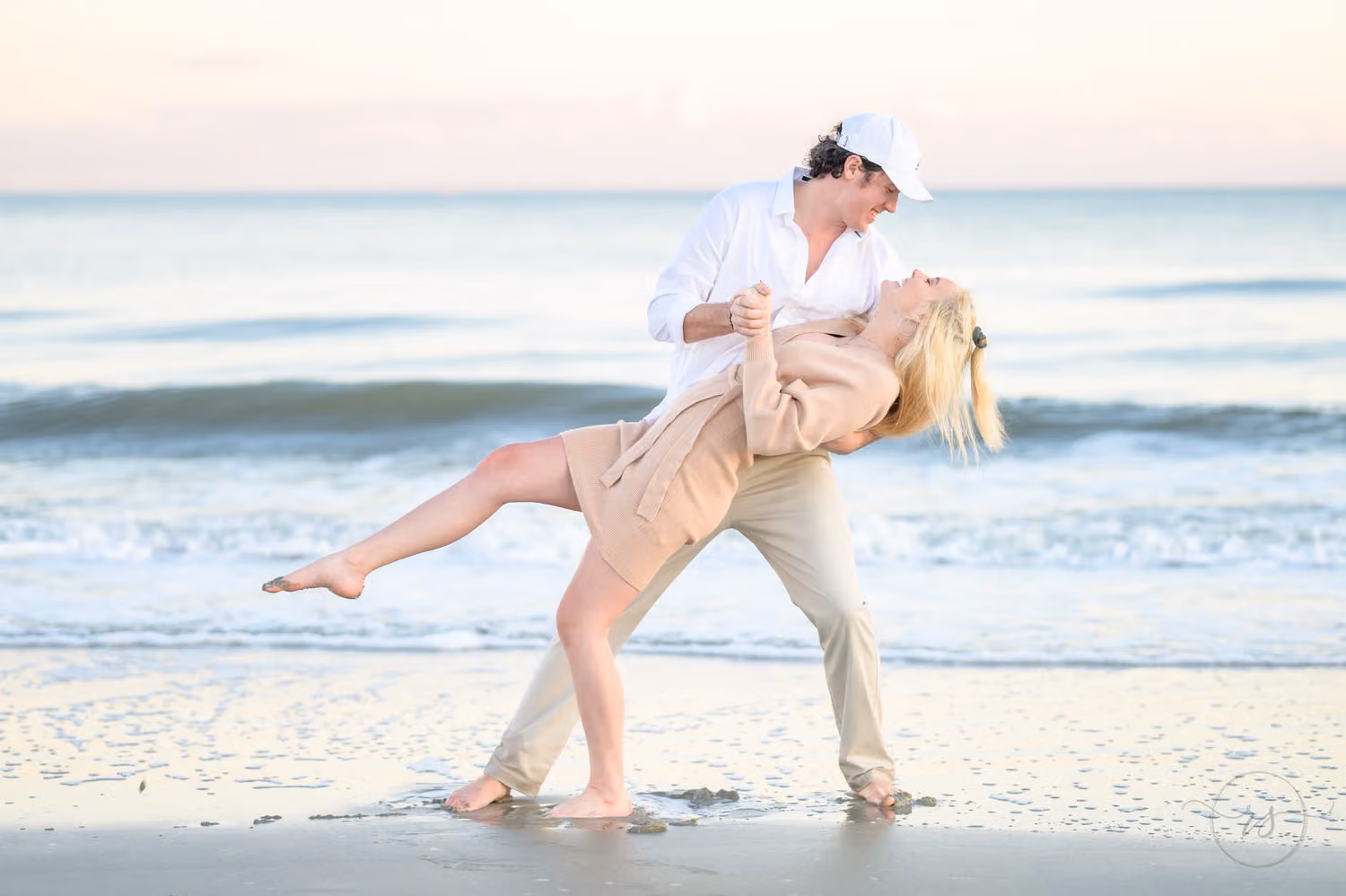 Man in a white cap dipping his fiancée back as she kicks up a leg by the ocean at Huntington Beach State Park