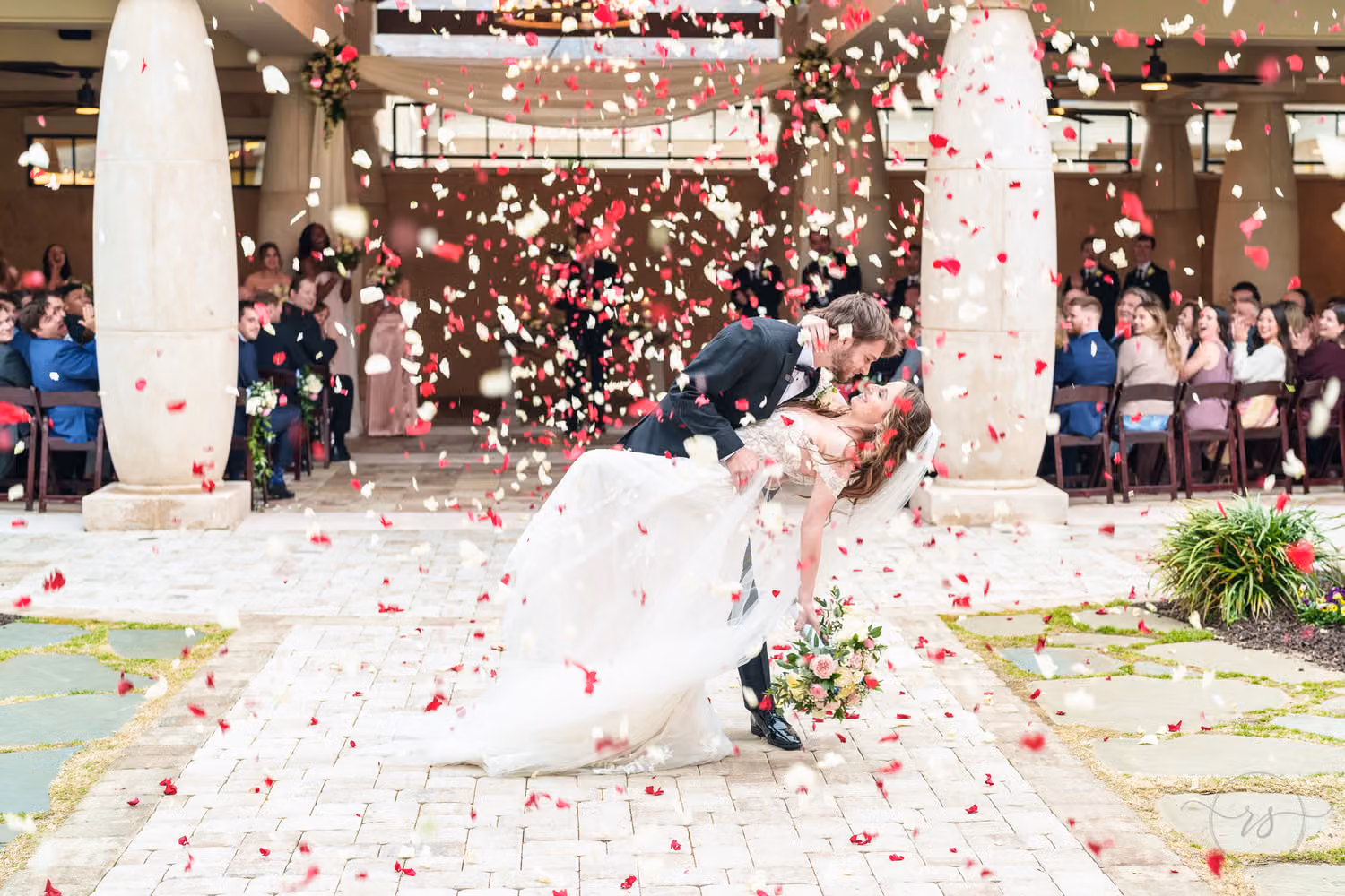 Groom dipping the bride as guests toss red and white flower petals after the ceremony at 21 Main Events