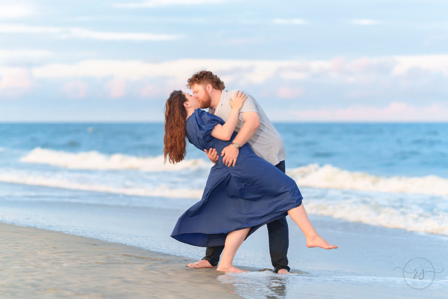 Man dipping his fiancée in a blue dress for a kiss by the waves under puffy white clouds at Huntington Beach State Park
