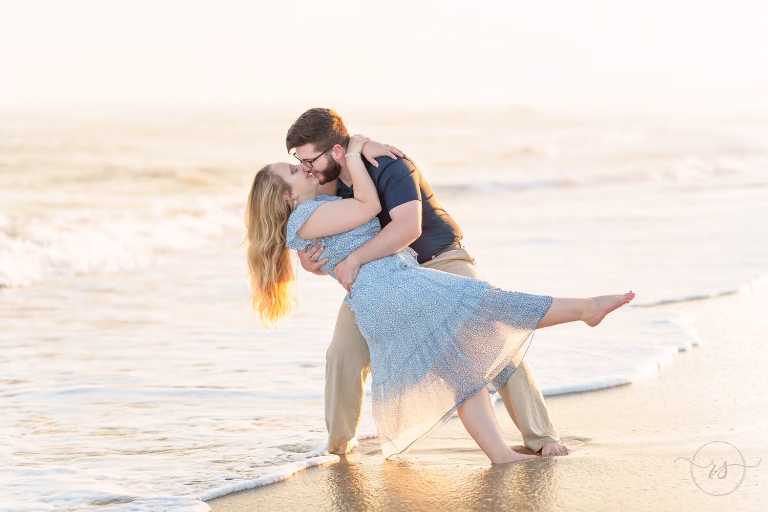 Man dipping his fiancée in a light blue dress as she kicks up a leg by the waves at Huntington Beach State Park