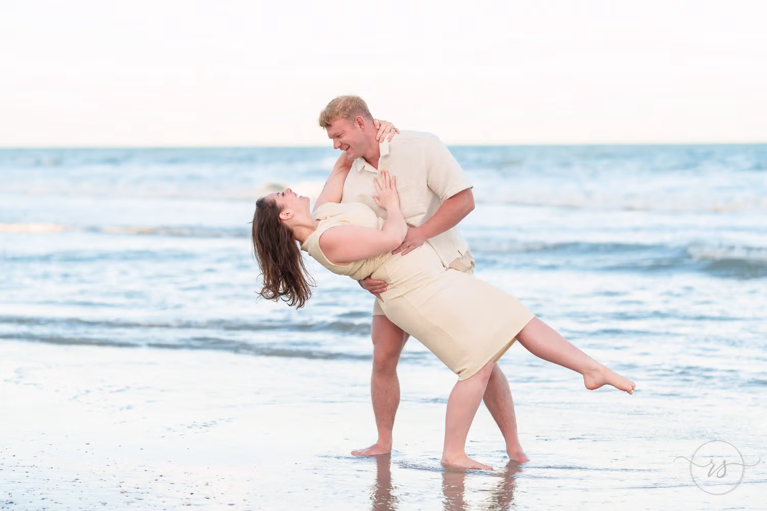 Barefoot woman kicking up her leg as her fiancé dips her for a kiss in the shallow water at Seawatch Resort