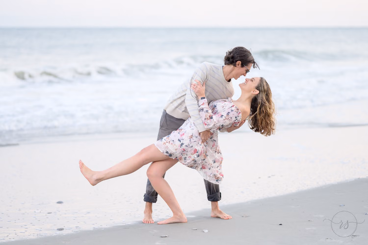 Man dipping his barefoot fiancée in a floral dress as they kiss by the waves at the Marriott in Myrtle Beach