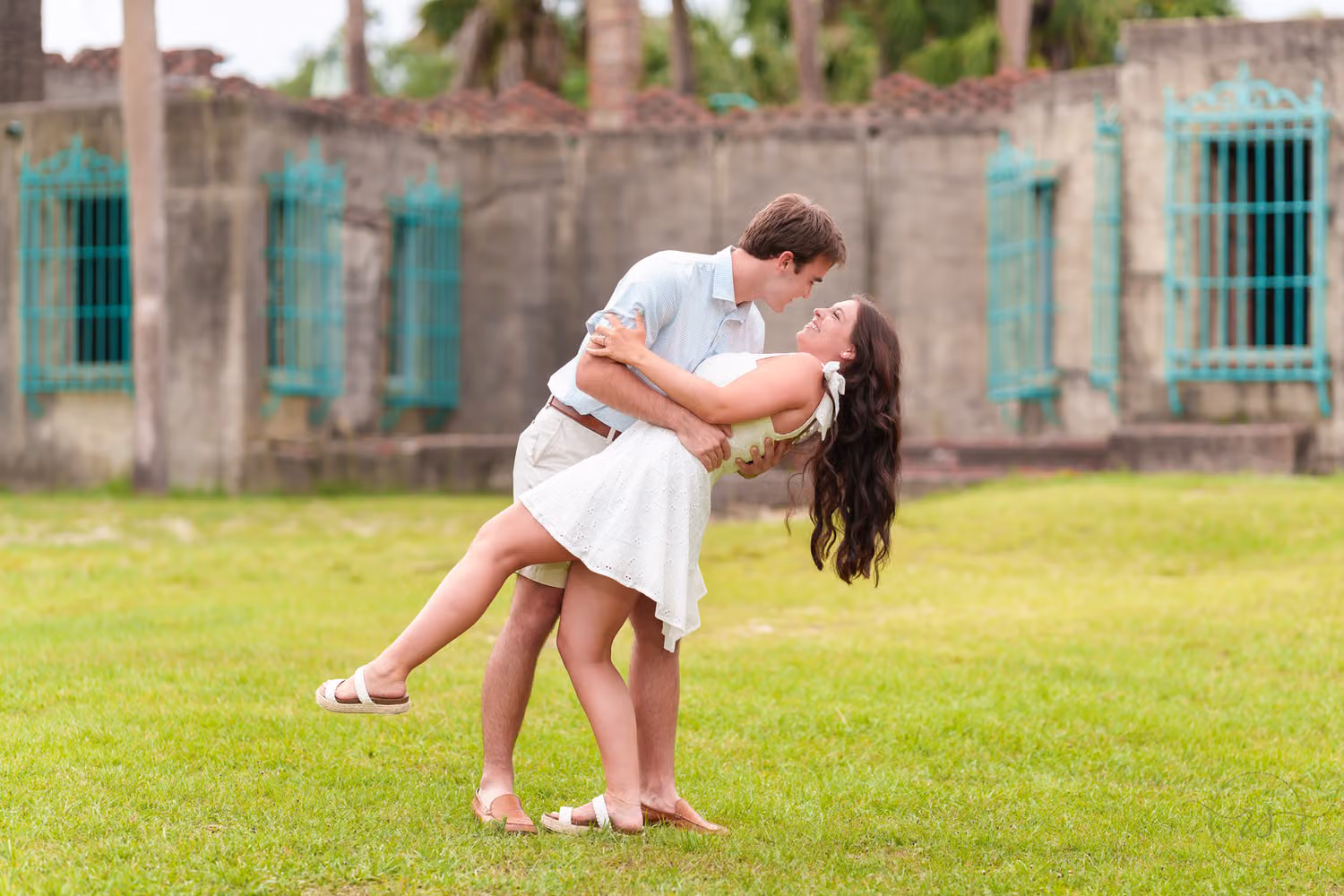 Man dipping his fiancée for a kiss on the grass by the teal windows of the Atalaya Castle at Huntington Beach State Park