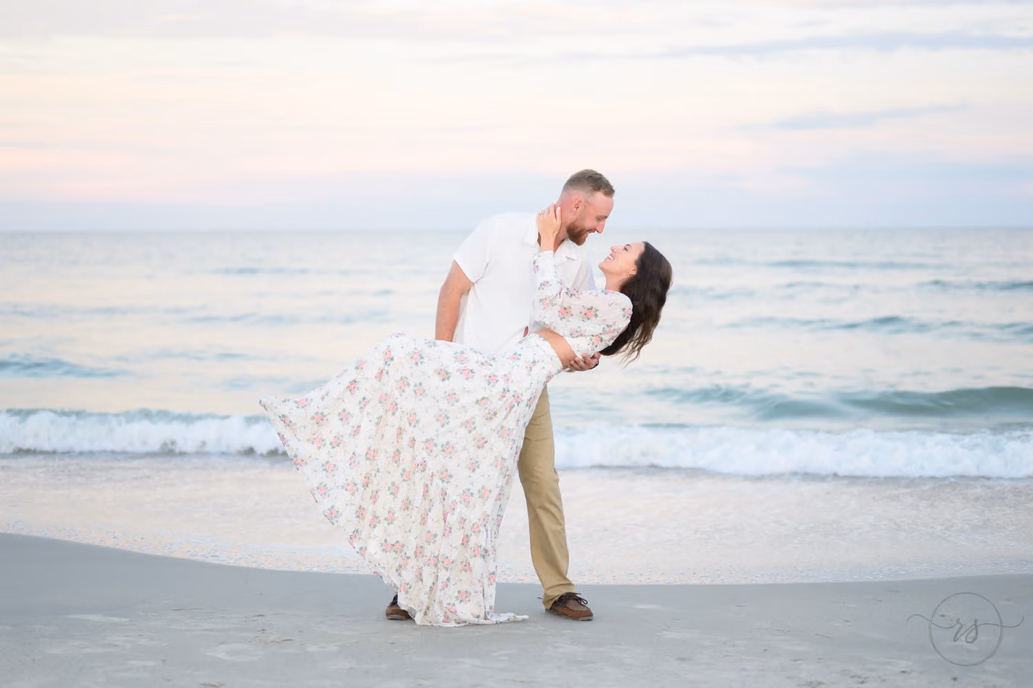 Man dipping his fiancée in a long floral dress at the water's edge in pastel evening light at Huntington Beach State Park