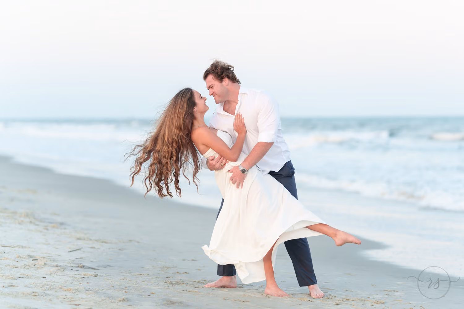 Man dipping his fiancée as her long hair swings out over the wet sand by the waves at Huntington Beach State Park
