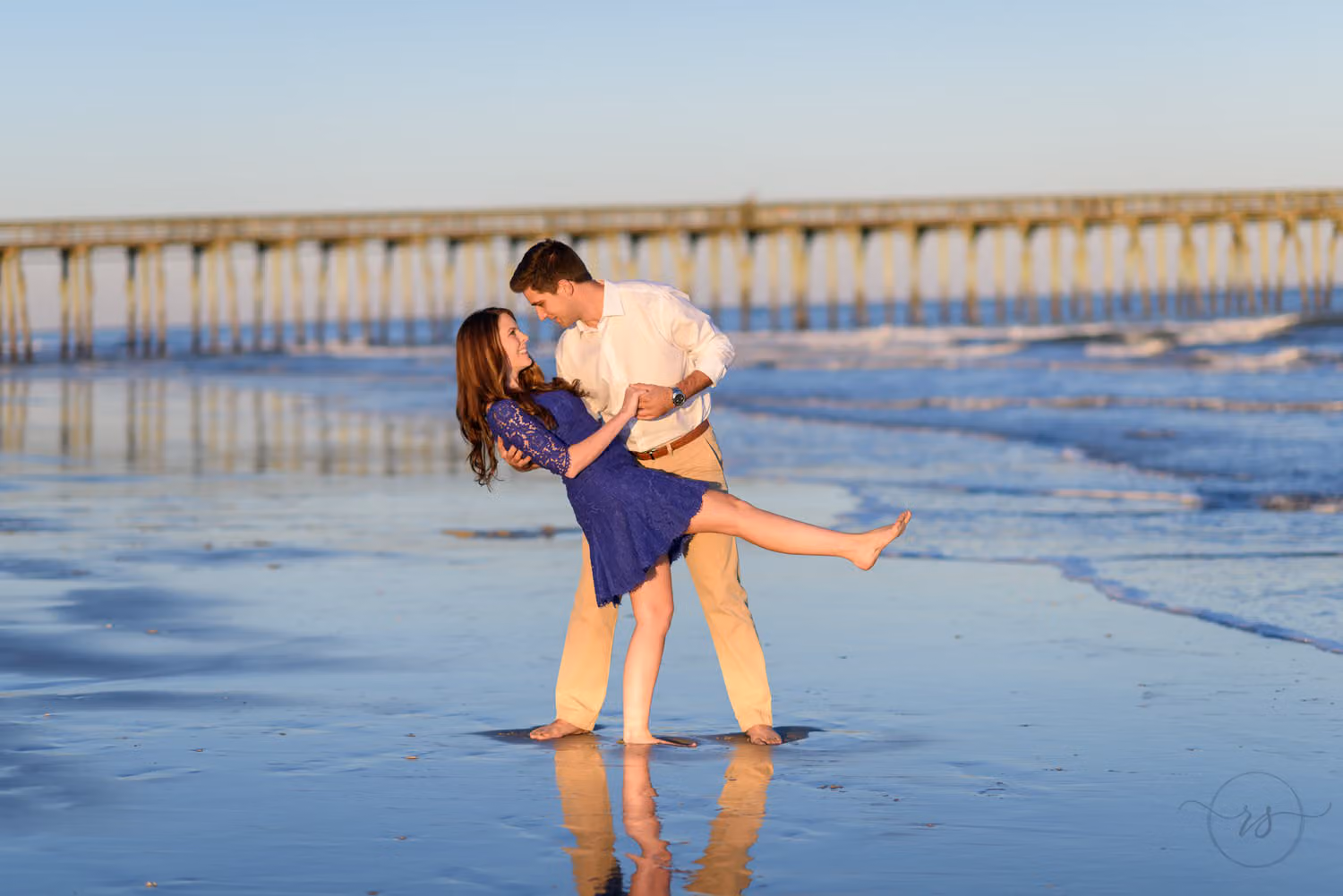 Man dipping his fiancée in a blue dress with their reflection in the wet sand and the pier behind at Myrtle Beach State Park