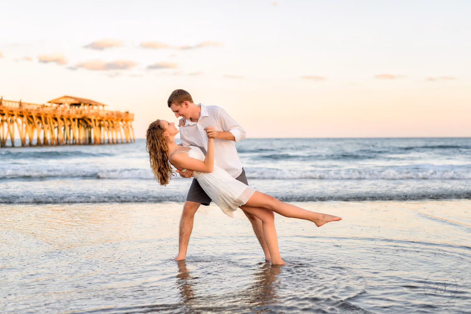 Man dipping his fiancée as she kicks up a leg in the shallow water with the pier behind at sunset at Myrtle Beach State Park