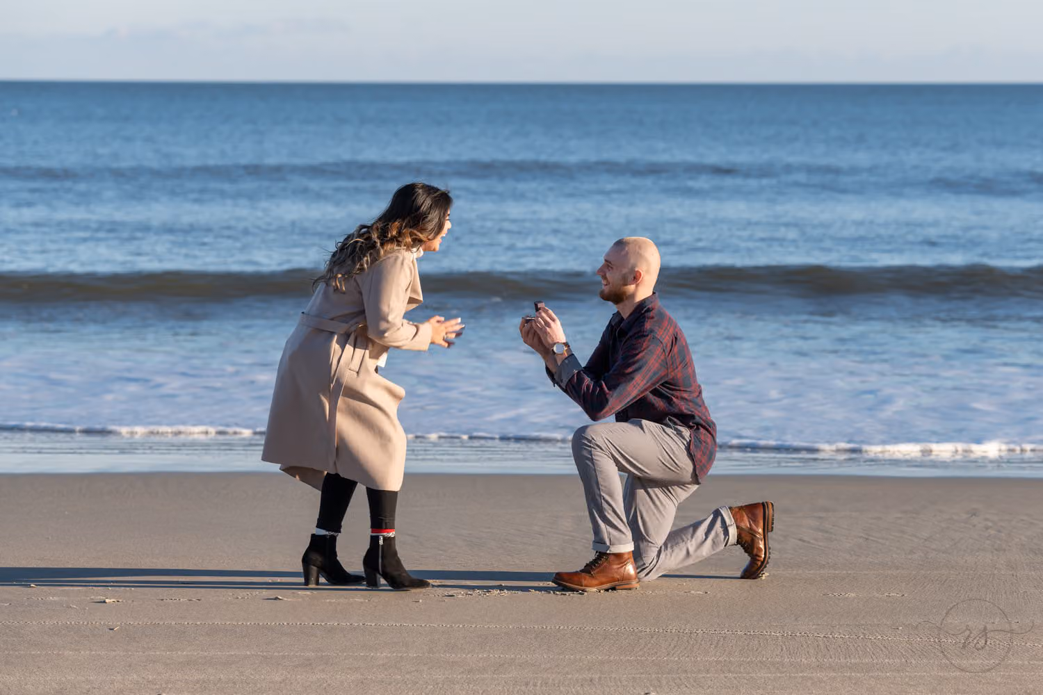 Man on one knee proposing as his girlfriend in a long coat leans in surprised on the beach at Huntington Beach State Park